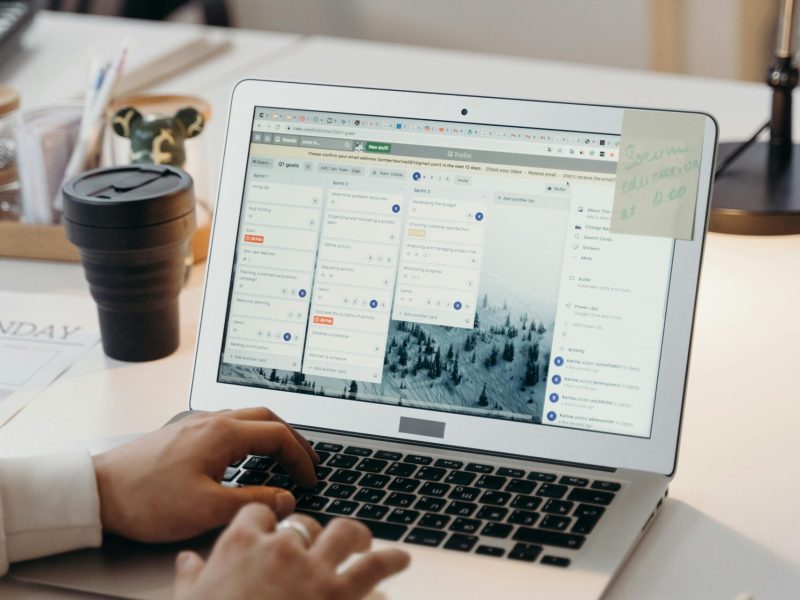 Hands typing on a laptop in a modern office setting with notes and task management software visible on the screen.